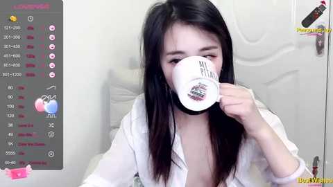 Media: Video of a young Asian woman with long black hair, wearing a white robe, drinking from a mug with \"My Pillow\" printed on it, in a minimalist bedroom.