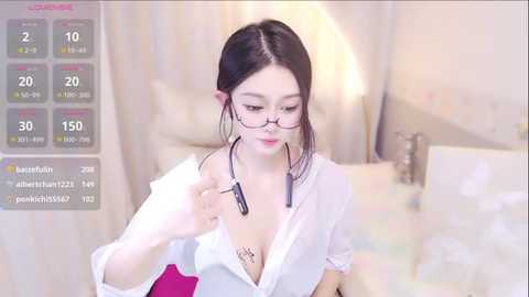 Media: Video of an East Asian woman with fair skin, long black hair, and glasses, wearing a white shirt, sitting on a bed with white sheets, looking at a smartphone displaying health data.