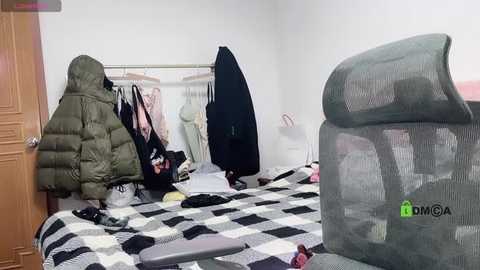 Media: Video of a messy bedroom with a checkered bedspread, clothes scattered, a green puffer jacket, a black jacket, and a grey armchair.