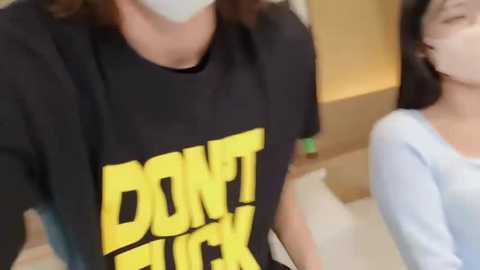 Media: Video of two people: a person in a black \"DON'T F**K WITH ME\" shirt, and another in a white top, standing indoors with blurred background.