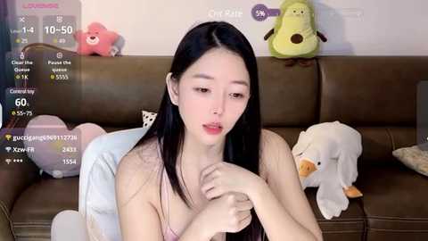 Media: Video of an East Asian woman with long black hair, light skin, and small breasts, sitting on a brown couch surrounded by plush toys, including a duck and a frog.