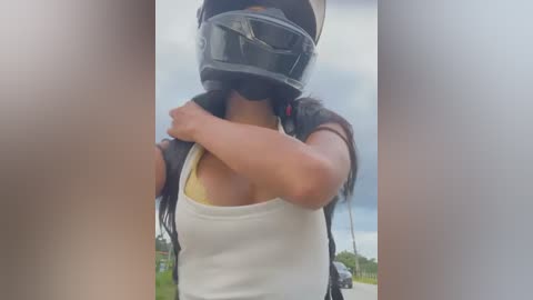 Media: A video of a woman wearing a black motorcycle helmet and a white tank top, adjusting the strap with her right arm. Background shows a cloudy sky and distant greenery.