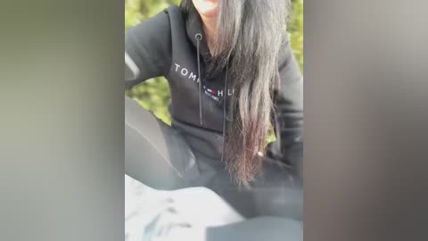 Media: A video of a young woman with long, straight black hair, wearing a black hoodie with \"Tommy Hilfiger\" in white on the chest, and black leggings. She's sitting on a stone surface outdoors, surrounded by greenery.