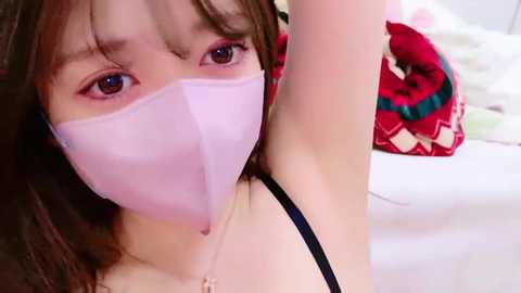 Media: Video of an Asian woman with fair skin, wearing a pink face mask, black bra, and a colorful patterned blanket in the background.