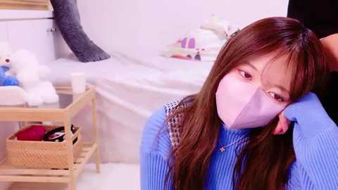 Media: Video of a young Asian woman with long brown hair wearing a blue sweater, a pink face mask, and a white headband, sitting in a cozy, well-lit room with toys, a teddy bear, and a basket.