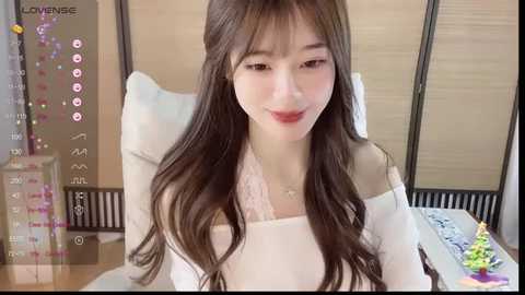 A video of a young Asian woman with long, wavy brown hair, fair skin, and light makeup. She wears a white off-shoulder top and sits at a wooden desk with a calendar, colorful decorations, and a cake.