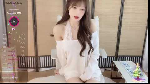 Media: Video of an Asian woman with long black hair, fair skin, and red lipstick, wearing a white off-shoulder blouse, seated on a wooden floor in a minimalist room with a beige screen.