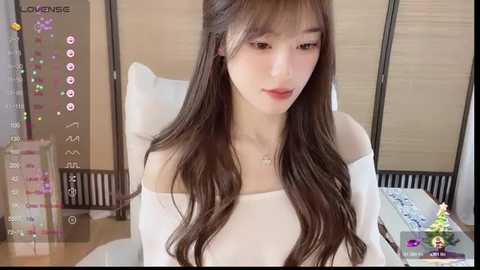 A video of a young Asian woman with long, wavy brown hair, wearing a white off-shoulder top, sitting indoors. The background features a wooden wall, a bed, and a table with a smartphone displaying a camera app.