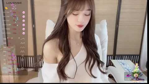 A video of a young Asian woman with long, wavy brown hair, wearing a white off-shoulder top, sitting on a white chair in a minimalist room with wooden paneling and a clock.