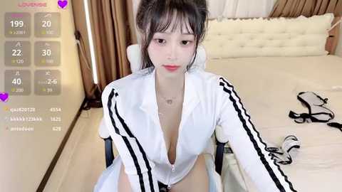 Media: A video of an Asian woman with fair skin and long, black hair in a white, open-front tracksuit, sitting on a white chair in a modern bedroom.