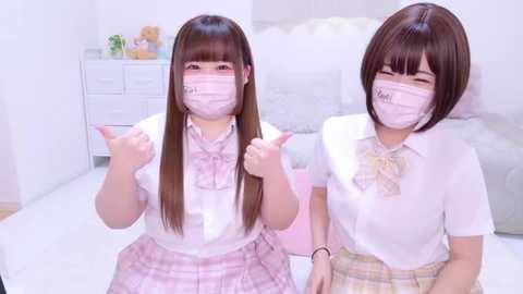 Media: Video of two young Asian girls with long brown hair, wearing white shirts, pink plaid skirts, and face masks, giving thumbs-up, in a white room with a bed and dresser.