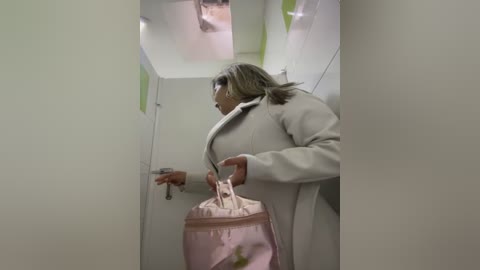 Media: A video of a woman with shoulder-length blonde hair, wearing a white coat and pink pants, holding a pink handbag, standing in a narrow, white-walled, reflective hallway.