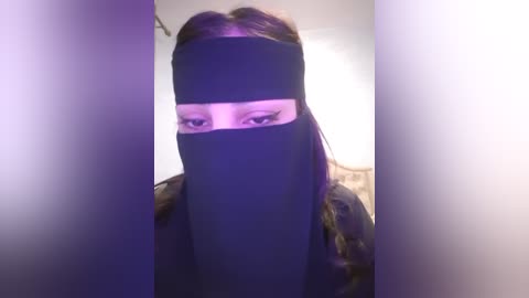 Media: A video of a person with a black face mask covering their eyes, wearing a dark coat, taken indoors with a blurred background.