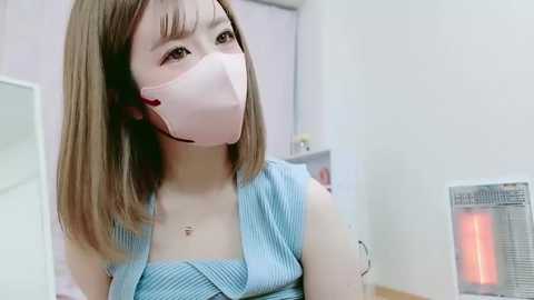 Media: Video of an Asian woman with shoulder-length brown hair wearing a blue striped dress, a pink face mask, and a necklace, standing indoors beside a heater and a white wall.