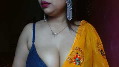 Media: Video of a South Asian woman with medium skin tone, wearing a blue bra and an orange sari with colorful floral embroidery. She has long, straight black hair and wears large, silver, beaded earrings.