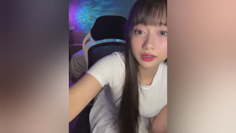 Media: Video of an Asian woman with long black hair and fair skin, wearing a white shirt, sitting in a black gaming chair under a colorful LED light.