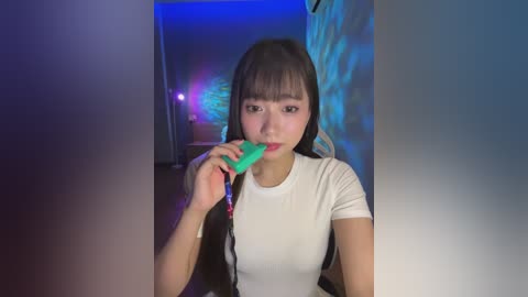 Media: Video of an East Asian woman with straight black hair and fair skin, wearing a white t-shirt, sucking a green lollipop. The background features a dimly lit room with blue and purple lighting.