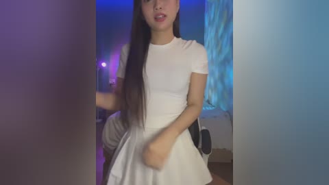 Media: Video of a young Asian woman with long black hair, wearing a white short-sleeved dress, standing in a dimly lit room with blue and purple lighting, holding a white object.