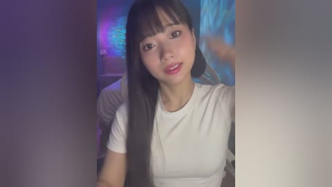 Media: Video of an East Asian woman with long black hair and fair skin, wearing a white T-shirt, smiling softly. The background features a blurred, blue light.