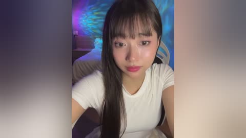 Media: Video of a young Asian woman with long black hair, wearing a white T-shirt, sitting indoors with a blue wall and a nightstand in the background.