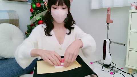 Media: A video of a young woman with light skin, wearing a white fluffy cardigan, pink face mask, and a Christmas-themed headband, seated at a desk, crafting a DIY project with a hot glue gun.