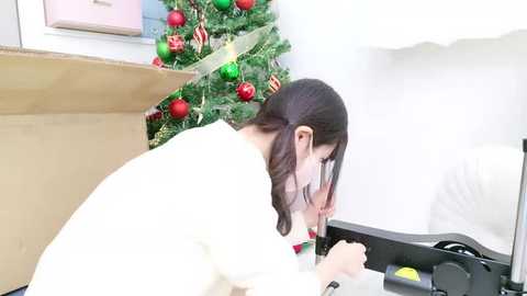 Media: Video of an Asian woman with long dark hair, wearing a white sweater, leaning over a desk, unpacking a box of Christmas decorations. A decorated Christmas tree is visible in the background.