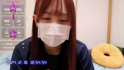 Media: A video shows an Asian woman with long, straight auburn hair, wearing a white face mask, black jacket, and lying on a bed. The background features a yellow pillow and a white wall.