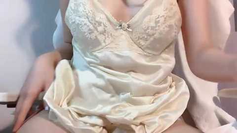 Video of a woman in a satin, cream-colored lingerie set with lace detailing, seated with one hand on her thigh, revealing a hint of cleavage.