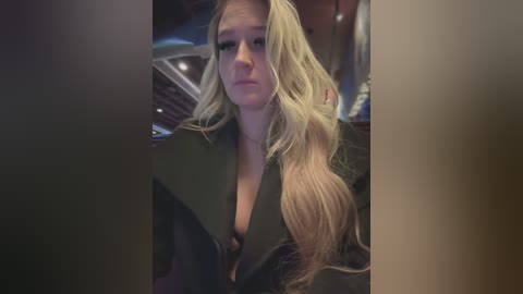 Media: Video of a blonde woman with long hair, wearing a black leather jacket, sitting in a dimly lit room, with blurred objects in the background.