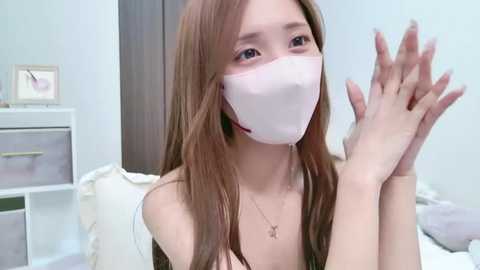 Media: Video of an Asian woman with long brown hair, wearing a white face mask and a black off-shoulder top, clapping her hands, in a modern bedroom with light walls and white furniture.