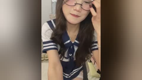 Media: A video of an Asian woman with long brown hair and glasses, wearing a navy and white sailor-style shirt, adjusting her glasses in a dimly lit room with a beige wall and a bed visible in the background.