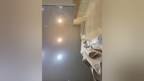 Media: Video of a narrow, modern bathroom with a tiled shower on the left, a white towel rack with neatly folded towels on the right, and soft, ambient lighting creating a warm, cozy atmosphere.