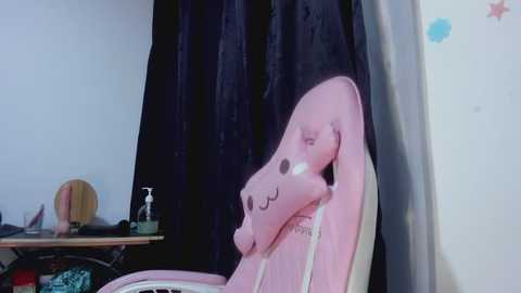 Media: Video of a pink, plush chair with a smiling face, in a room with a black curtain, white wall, and a wooden shelf holding a mirror, soap, and bottle.