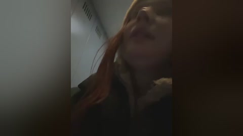 Media: A dimly lit, blurred video showing a woman with long, red hair, wearing a dark jacket, standing in a narrow, dimly lit hallway with white walls and a faint light source.