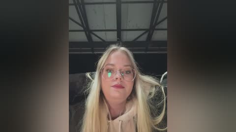 Media: Video of a blonde woman with glasses and a light tan jacket, sitting in an industrial-looking room with metal beams overhead.