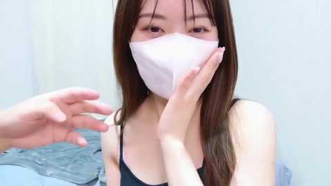 Media: Video of an East Asian woman with long brown hair, wearing a black spaghetti-strap top and white surgical mask, hands placed near her mouth, in a simple, indoor setting with light-colored walls.