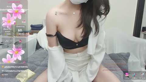 Media: Video of an Asian woman with fair skin, wearing a black bra, white robe, and face mask, sitting on a gray bed, with floral filters overlaying the image.