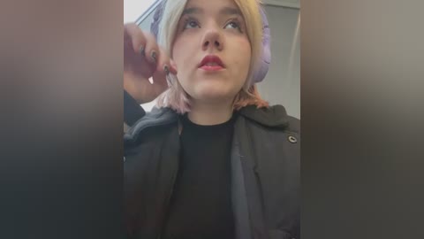 Media: Video of a young woman with light skin and pinkish-blonde hair, wearing large purple headphones, a black jacket, and a black top, looking upwards with a contemplative expression.