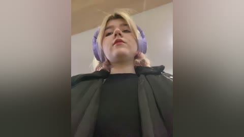 Media: A video of a young Caucasian woman with light skin and blonde hair, wearing purple headphones, a black top, and a grey jacket, standing in a dimly lit room with a beige wall and a wooden ceiling.