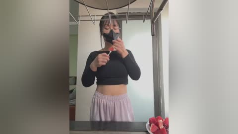 Media: A video of a young Asian woman with light skin, wearing a black face mask and a black long-sleeved crop top, taking a mirror selfie in a dimly lit bathroom. She holds a phone in her right hand.