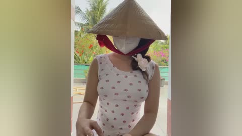 Media: Video of an Asian woman in a white sleeveless shirt with small red flower prints, wearing a traditional conical hat and face mask, standing outdoors with a lush garden background.