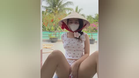 Media: Video of an Asian woman in a floral-patterned white top and white panties, wearing a conical hat and face mask, sitting with legs spread on a balcony overlooking lush greenery.