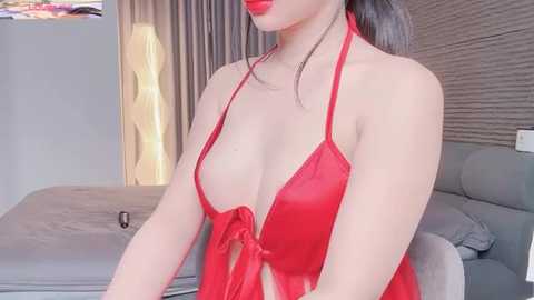 Video of a fair-skinned woman in a revealing red satin halter dress, wearing red lipstick, standing in a modern bedroom with gray walls, a bed, and a standing lamp.