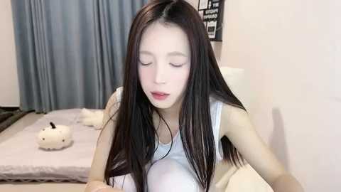 Media: A video of a young Asian woman with long, straight black hair, wearing a white tank top, lying on a bed with light-colored sheets and pillows. The room has a simple, neutral decor with a gray curtain in the background.