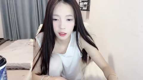 Media: Video of an East Asian woman with long black hair, fair skin, and light makeup, wearing a white sleeveless top, lying on a bed with light gray sheets, in a softly lit room.