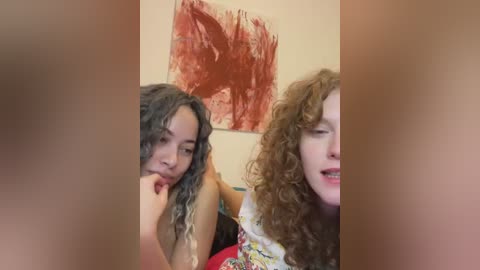 Media: Video of two young women with curly hair, one with dark hair and the other with light brown hair, sitting side by side, smiling. Background features a red abstract painting on a beige wall.