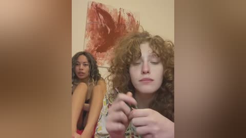 Media: Video of two women in a cozy room. One with curly brown hair, wearing a floral dress, holds a phone; the other, with long, wavy dark hair, is topless, leaning against a wall with abstract art.