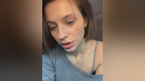 Media: Video of a young Caucasian woman with straight brown hair, light skin, and a delicate face, wearing a light blue off-shoulder top, looking introspective.