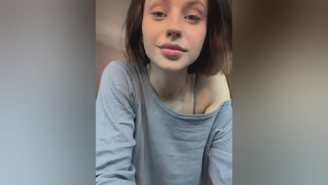 Media: Video of a young Caucasian woman with fair skin, straight shoulder-length brown hair, wearing a loose gray off-shoulder top, standing indoors with blurred background.