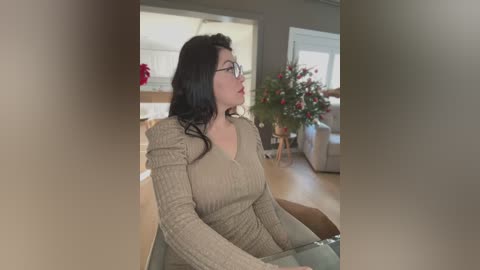 Video of a woman with long black hair, wearing glasses, a beige sweater, and light pants, sitting indoors, facing a floral arrangement.